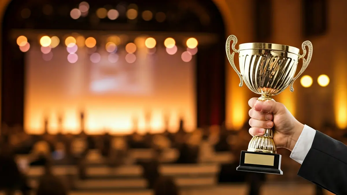 Image of a hand holding a trophy at an awards ceremony, with blurred figures in the background.